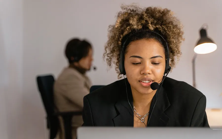 A support engineer speaking through a headset at a desk, a colleague working behind.